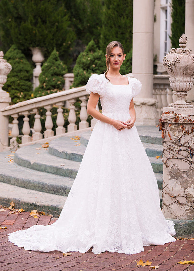 Woman in a white wedding dress standing on a brick path with stone columns and greenery in the background.