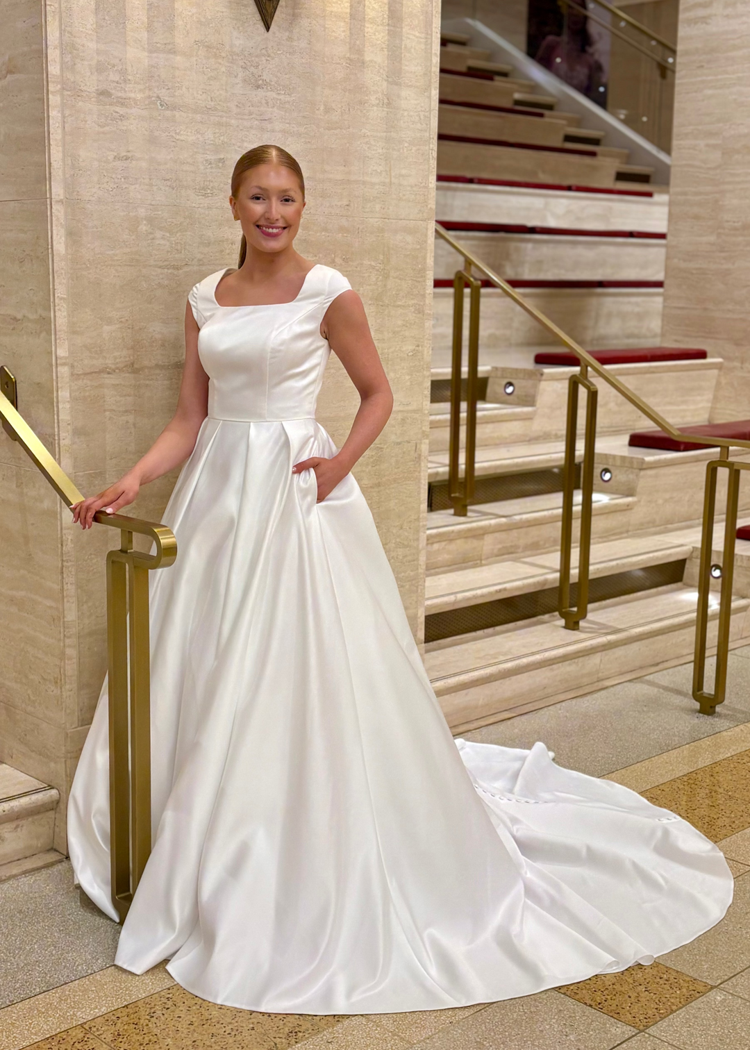 Woman in a white modest wedding dress standing in a grand hallway with marble walls and stairs.