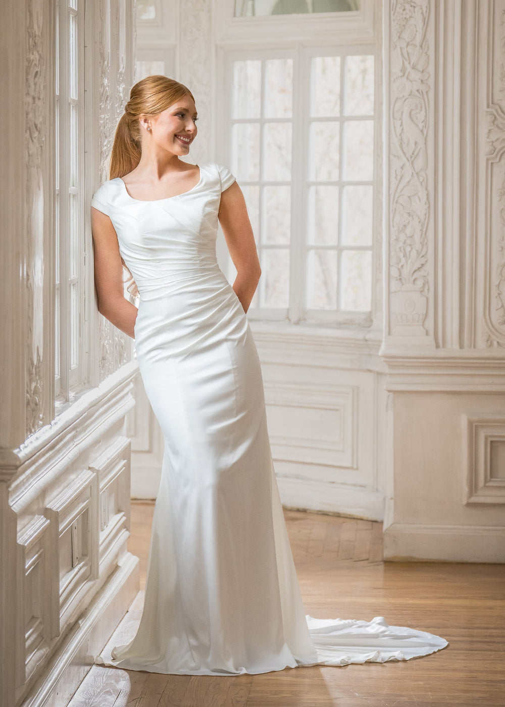 Woman in a white wedding dress standing in an elegant room with high ceilings and large windows.