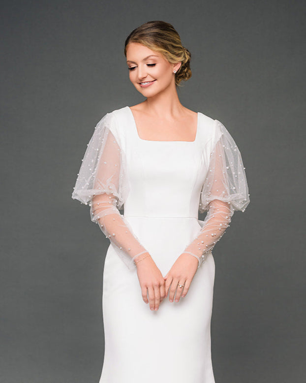 Woman wearing an ivory modest wedding dress with a sheer tulle and pearl sleeves on a gray background