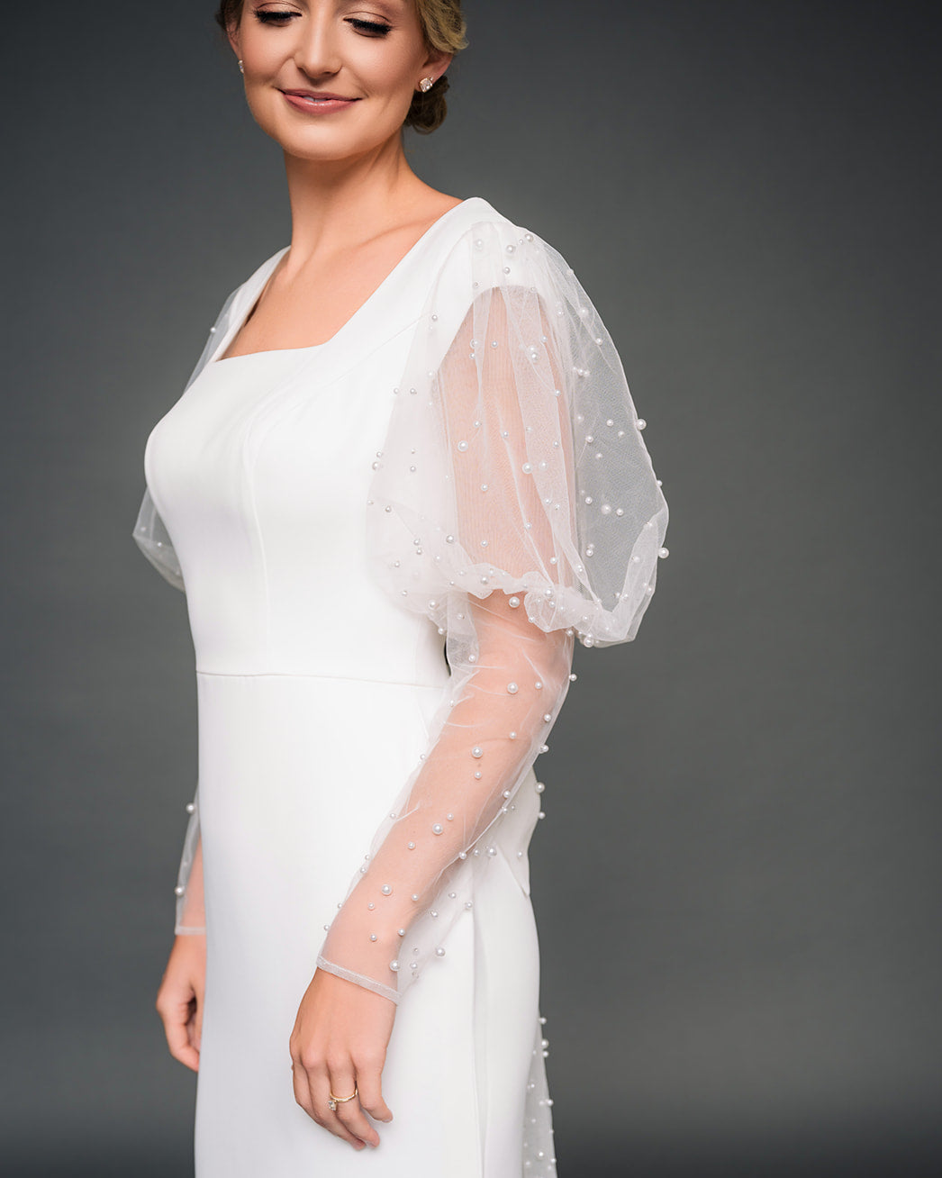 Woman wearing a modest ivory dress with a sheer, pearl tulle against a dark background